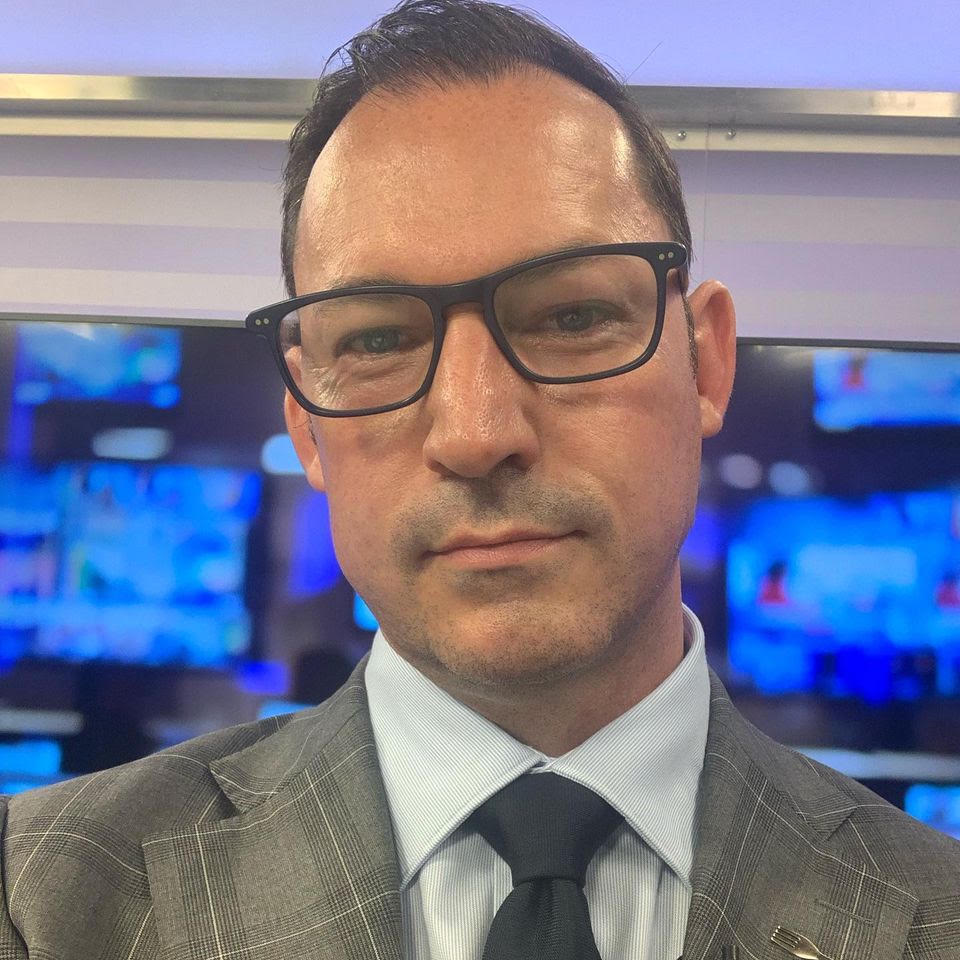 Wes Lambert wearing glasses and a grey suit, with blue screens behind him
